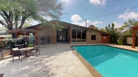 This community pool facility offers a well-designed outdoor retreat centered around a large, sparkling swimming pool with a generously sized concrete deck. The deck is equipped with numerous lounge chairs and patio umbrellas, providing ample space for sunbathing and relaxation. A standout feature is the wooden pergola positioned at one end of the pool, offering a shaded seating area framed by mature trees and an open sky backdrop.

The single-story clubhouse building anchors the space architecturally, featuring striking floor-to-ceiling glass windows and warm wood-accented framing that creates a mid-century modern aesthetic. The building's interior connects directly to the pool deck via glass sliding doors, allowing for seamless indoor-outdoor flow and clear sightlines to the pool. A covered porch area with traditional wood-framed doors serves as a welcoming entrance, complete with posted pool rules and a wall-mounted light fixture.

The pool area is enclosed by a black iron security fence with a gated entry, ensuring a safe and private environment. Lush, mature trees surround the perimeter, providing natural shade and a park-like atmosphere. The overall ambiance is open and airy, with abundant natural light and a bright, resort-style feel suited for community leisure and recreation.