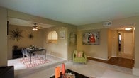 This single-floor residence offers an open and interconnected layout designed for comfortable everyday living. The living area is carpeted and bathed in a warm, neutral color palette, with walls accented by decorative artwork that adds a touch of personality to the space. A pass-through archway connects the living and dining zones to the kitchen beyond, promoting an open flow while maintaining defined areas for each function. The dining area features a ceiling fan with an integrated light fixture, providing both comfort and illumination. The kitchen is partially visible through the arched pass-through, hinting at a practical and accessible layout. Throughout the space, textured ceilings add architectural character, while HVAC vents integrated into the walls and ceilings ensure year-round climate control. Large baseboards frame the carpeted floors, contributing to the clean, finished look of the interior. The overall ambiance is casual and inviting, with a cohesive design that balances functionality with a lived-in warmth, making it a versatile and approachable single-level living space.