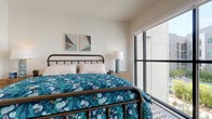 The Cascade is a thoughtfully designed single-floor residence that blends modern aesthetics with functional living. The primary bedroom serves as the heart of the space, featuring an iron-frame bed, light wood-style flooring, and a patterned area rug that adds visual warmth. A large floor-to-ceiling window floods the room with natural light while offering views of the surrounding community. A ceiling fan with recessed lighting ensures year-round comfort. The open layout flows seamlessly into a living area equipped with a flat-screen television atop a rustic wood media console, a compact kitchenette with gray cabinetry, white countertops, and small appliances — ideal for convenient, everyday use. A full-length mirror and coat rack station near the hallway provide practical organization. Clean, white walls throughout create a bright, airy ambiance. The bathroom is finished with classic white subway tile extending from floor to mid-wall, wood-look flooring, a tub-shower combination with a curtain rod, and accessibility grab bars for added safety. Curated artwork throughout the space adds personality and a contemporary flair, making the Cascade a comfortable and stylish single-level retreat.