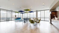 This stylish high-rise amenity lounge is a well-appointed, single-floor communal space designed for both socializing and relaxation. Floor-to-ceiling glass walls wrap around the main area, flooding the interior with natural light and offering sweeping panoramic views of the surrounding cityscape. The open layout is thoughtfully organized into distinct seating zones, including a lounge area anchored by a vibrant teal sofa, mid-century-inspired swivel chairs, and a blue area rug atop polished concrete flooring. Dining tables with upholstered green and grey chairs provide additional gathering spots. Overhead, bold geometric acoustic panels in blue and green hues serve as striking suspended ceiling installations, complementing the space's contemporary aesthetic. A dedicated kitchenette area features dark wood cabinetry, a patterned geometric tile backsplash, under-counter appliances, and ample counter space, making it functional for events and gatherings. An eye-catching mural with a nature-inspired motif adds an artistic focal point along one wall. The space also connects to an outdoor terrace, further enhancing the sense of openness. The overall design balances energetic color with clean, modern lines, creating a dynamic and inviting environment well-suited for community gatherings and everyday use.
