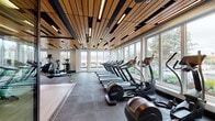Located at 522–538 San Antonio Rd in Mountain View, California, this contemporary fitness center is thoughtfully designed to support a full range of workout modalities in a single, well-appointed space. The facility is defined by its striking wood-slat ceiling with integrated linear lighting, which adds warmth and sophistication throughout, complemented by large floor-to-ceiling windows that flood the interior with natural light and offer views of the surrounding greenery.

The cardio zone lines the window-facing perimeter, housing a generous row of treadmills, ellipticals, and stationary bikes — all positioned to take advantage of the outdoor views. A dedicated cycling and rowing studio features a light wood floor, wall-to-wall mirrors, and sliding glass doors that connect it seamlessly to the main floor, creating a flexible, open-concept layout.

The strength and functional training area is equally well-equipped, offering a comprehensive free weights section with dumbbells, kettlebells, medicine balls, BOSU trainers, resistance bands, and TRX suspension systems. A power rack, cable machines, and adjustable benches round out the strength equipment offerings. Punching bags and a water fountain station are also present, adding further versatility. Large-format tile flooring and rubber matting provide a durable, cohesive finish throughout the space.