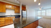 This single-floor residence offers a well-designed open layout that seamlessly connects the kitchen and living areas. The kitchen is a standout feature, showcasing rich honey-toned wood cabinetry that extends to the upper walls, providing generous storage throughout. Dark granite countertops run along the perimeter and across a substantial center island, which is illuminated by pendant lighting and equipped with built-in electrical outlets. Stainless steel appliances, including a refrigerator, range, microwave, and dishwasher, complement the space, while track lighting overhead ensures thorough illumination. Light ceramic tile flooring anchors the kitchen and transitions into carpeted areas in the living room and bedroom. The living area benefits from natural light through large windows fitted with horizontal blinds, and a ceiling fan adds comfort. The bedroom is quietly spacious, with soft carpeting, a ceiling fan, and dual windows that invite daylight. The bathroom is finished with light tile flooring, wood cabinetry for storage, and connects conveniently to a walk-in closet and an in-unit laundry area. Overall, the space presents a functional and cohesive design with consistent warm-toned finishes throughout.