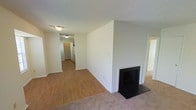 This single-story space offers a practical and functional layout with notable design details throughout. The main living area is carpeted and centers around a black-surround fireplace set into a clean white wall, serving as a natural focal point for the room. A textured ceiling adds dimension overhead, while flush-mounted lighting provides consistent illumination. Neutral white walls throughout keep the space feeling bright and versatile.

A sliding glass door, dressed with vertical blinds, floods the carpeted living space with natural light and provides access to an outdoor area. The transition between rooms is marked by a shift in flooring, moving from plush carpet in the living area to hardwood-style flooring in the adjoining hallway and additional spaces beyond. This corridor connects to further rooms within the unit, visible in the background.

The overall aesthetic is clean and understated, with a neutral color palette of whites and beiges that creates a blank canvas adaptable to a variety of furnishing styles. The open feel of the living area, combined with natural light from the sliding door, contributes to an airy and welcoming ambiance throughout the space.