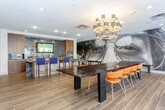 This contemporary single-floor space is designed for both entertaining and gathering, combining bold artistic elements with practical amenities. The open layout is anchored by wood-look tile flooring throughout, creating a warm and cohesive aesthetic across the entire area.

The dining area is a standout feature, centered around a large, dark-topped communal table lined with vibrant orange chairs. An eye-catching black-and-white oversized mural depicting a close-up face with sunglasses commands one wall, adding a striking artistic focal point. A sculptural multi-shade chandelier hangs above the table, providing ambient lighting with a decorative edge. Multiple floor-to-ceiling sliding glass doors run along one side, flooding the space with natural light and providing seamless access to an outdoor patio area.

Adjacent to the dining space is a well-appointed kitchen, equipped with stainless steel appliances including double wall ovens, a microwave, and a sink. Rich wood cabinetry lines the walls above a metallic tile backsplash, and a wall-mounted television is centrally positioned. A granite-topped island with blue upholstered barstools serves as a casual seating and workspace area, making this space as functional as it is visually engaging.