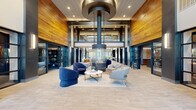 This impressive amenity facility at Acero Southtown in San Antonio spans a generously proportioned single floor, offering a comprehensive suite of community spaces designed for both relaxation and activity. The centerpiece is a dramatic double-height lobby lounge featuring soaring ceilings clad in warm wood paneling, bold steel-framed architectural columns in deep navy, and a striking suspended cylindrical fireplace that anchors a curated seating arrangement of blue and grey upholstered chairs. Large grid-pane windows flood the space with natural light, enhancing the open, airy ambiance. Adjacent to the lobby, a full community kitchen impresses with rich walnut cabinetry, sleek grey quartz countertops, a wood-paneled ceiling, and ample workspace ideal for entertaining. The dining area features a substantial live-edge communal table beneath pendant lighting, with exposed brick accents and the Acero Southtown brand prominently displayed. A dedicated game lounge with textured brick walls, curated wall art, and a foosball table offers a casual social retreat. The fitness facilities include a well-equipped strength and cardio exercise room with a wood-planked ceiling, plus a separate yoga and studio room with floor-to-ceiling mirrors, built-in shelving, and an integrated display screen. Multiple hallways connect the spaces seamlessly throughout.