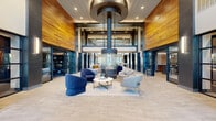 This impressive amenity facility at Acero Southtown in San Antonio spans a generously proportioned single floor, offering a comprehensive suite of community spaces designed for both relaxation and activity. The centerpiece is a dramatic double-height lobby lounge featuring soaring ceilings clad in warm wood paneling, bold steel-framed architectural columns in deep navy, and a striking suspended cylindrical fireplace that anchors a curated seating arrangement of blue and grey upholstered chairs. Large grid-pane windows flood the space with natural light, enhancing the open, airy ambiance. Adjacent to the lobby, a full community kitchen impresses with rich walnut cabinetry, sleek grey quartz countertops, a wood-paneled ceiling, and ample workspace ideal for entertaining. The dining area features a substantial live-edge communal table beneath pendant lighting, with exposed brick accents and the Acero Southtown brand prominently displayed. A dedicated game lounge with textured brick walls, curated wall art, and a foosball table offers a casual social retreat. The fitness facilities include a well-equipped strength and cardio exercise room with a wood-planked ceiling, plus a separate yoga and studio room with floor-to-ceiling mirrors, built-in shelving, and an integrated display screen. Multiple hallways connect the spaces seamlessly throughout.