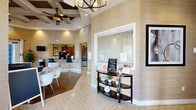 Located at Esplanade Park Circle in Orlando, FL, this single-floor community clubhouse offers a thoughtfully designed space that blends functionality with refined aesthetics. The entrance greets visitors with striking coffered ceilings adorned with dual ceiling fans and a chandelier, setting an immediately sophisticated tone. Warm, textured grasscloth-style wall coverings in neutral tones flow throughout, creating a cohesive and inviting atmosphere. The living room area features rich hardwood flooring, elegant French doors with floor-to-ceiling white drape panels, and a distinctive glass block window feature that allows natural light to fill the space. Comfortable seating, a wall-mounted television, and framed artwork complete the lounge ambiance. An adjoining workspace with desks and office seating provides a practical area for residents. The kitchen is well-appointed with warm wood cabinetry, dark granite countertops, stainless steel appliances including a refrigerator, microwave, and dishwasher, complemented by cork-style tile flooring. Additional interior doors lead to private rooms, and a welcoming reception console near the entrance adds a communal, hospitality-forward touch to this polished clubhouse.