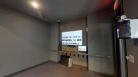 This single-level cycling studio is purpose-built for group fitness and individual training sessions. The space is anchored by a row of stationary spin bikes arranged on durable, low-pile carpeted flooring, providing a stable and comfortable surface for high-intensity workouts. Exposed spiral ductwork runs along the ceiling, lending the studio an open, industrial aesthetic that complements its modern, dark-toned interior palette of deep grays and charcoal.

A large wall-mounted flat-screen display is positioned at the front of the room, supported by a built-in wood-topped shelf and flanked by a secondary monitor, enabling instructors to lead virtual classes or stream workout content. Full-length wall mirrors span a significant portion of the studio, visually expanding the space and allowing participants to monitor their form during sessions.

Natural light enters through floor-to-ceiling sliding glass doors that open directly to an outdoor area, providing a refreshing connection to the exterior and easy ventilation. Recessed lighting supplements the natural light, ensuring the studio remains well-lit at all times. A dedicated entry door with an illuminated exit sign ensures safe and convenient access. The overall ambiance is sleek, focused, and energizing — well-suited for a modern fitness environment.