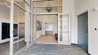 The Lower Lobby and Mailroom is a well-designed, functional entry space that combines modern aesthetics with practical amenities. The lobby is defined by its impressive double-height glass partition walls framed in sleek white metal, creating a sense of openness and allowing natural light to flow generously throughout. Large glass double doors at the street-facing entrance offer clear sightlines to the exterior streetscape, reinforcing the connection between indoors and out.

The flooring features a durable, patterned carpet in muted grey tones, providing a polished yet understated foundation throughout. A decorative pendant fan fixture suspended from the ceiling adds a subtle design accent overhead. Wall-mounted artwork, including a framed black-and-white clock print, contributes a touch of character to the otherwise clean, contemporary interior.

A stainless steel elevator serves as the functional centerpiece of the inner lobby, providing smooth access to upper floors. An adjacent seating bench offers a convenient waiting area for residents and guests. The mailroom area is equipped with an extensive array of individual black metal mailboxes mounted along the wall, accommodating a large number of residents with ease. The overall ambiance is bright, clean, and efficient — reflecting a thoughtfully designed space built to serve as a welcoming and organized gateway to the building.
