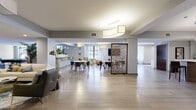 Located in the heart of Cleveland's vibrant East Sixth Street corridor, this single-floor amenity space offers a thoughtfully designed environment that blends modern aesthetics with functional comfort. Light wood-look tile flooring flows seamlessly throughout, unifying the various zones within the expansive open layout.

The central gathering area features multiple round white tables with contemporary seating, illuminated by sculptural pendant and drum lighting fixtures overhead. A decorative lattice-style room divider adds visual interest while subtly delineating spaces. Nearby, a lounge nook with large windows offers sweeping views of the Cleveland skyline, filling the area with natural light and providing a comfortable spot with upholstered seating and elegant accent decor.

A well-appointed kitchenette bar anchors one section of the space, complete with white cabinetry, a mosaic tile backsplash, quartz countertops, beverage refrigerators, a coffee station, and bar-height seating — ideal for casual gatherings. A textured white stacked-stone accent wall adds architectural warmth to an adjacent seating alcove furnished with sleek blue chairs.

Art-lined corridors connect the various areas, displaying a curated collection of framed works that lend a gallery-like character to the passageways. Cozy yellow wingback chairs and a wall-mounted flat-screen television create an inviting media nook. The overall ambiance is contemporary, polished, and welcoming.