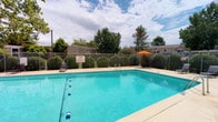 This amenity space features two distinct areas designed to support an active lifestyle. The outdoor pool area showcases a generously sized swimming pool with crystal-clear turquoise water, divided by a lane line and equipped with metal entry rails for easy access. The pool deck provides ample space for lounging, complemented by lounge chairs and a vibrant orange patio umbrella. The area is enclosed by a chain-link fence and bordered by neatly trimmed hedges and mature trees, creating a pleasant, park-like atmosphere open to the sky. Adjacent residential buildings are visible in the background, reinforcing the communal nature of the space.

The indoor fitness center features warm yellow walls accented by a bold orange and brown horizontal stripe, giving the room an energetic feel. Track lighting and wall sconces illuminate the space, which is finished with durable dark rubber flooring and a full-length wall mirror that enhances the sense of openness. Equipment includes a treadmill, elliptical machine, stationary bike, and a multi-function cable weight machine, offering a comprehensive range of cardiovascular and strength training options. A workout guide is posted on the wall for reference. Windows and a keyed exterior door provide natural light and direct outdoor access.