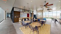Located at 2112 8th Street NW in Washington, DC's vibrant Shaw neighborhood, this single-floor communal space offers a thoughtfully designed environment that blends industrial architecture with warm, eclectic interior design. The open-plan layout is anchored by a striking lounge area featuring a built-in fireplace framed by floor-to-ceiling wood-paneled shelving with dark steel accents. A bold blue sectional sofa, a reclaimed wood coffee table, and a rich red area rug create an inviting focal point. A second lounge zone offers a low-profile red sectional against a wall of aged patina panels, complemented by adjustable industrial sconce lighting and layered textiles. The social and dining area features multiple round wood tables with metal chairs arranged across a jute rug, a bar-height counter with stools, and ceiling fans, with views of the surrounding cityscape visible through large windows. The fully equipped kitchen includes a central island with an induction cooktop, stainless steel appliances, a substantial range hood, subway tile backsplash, and slate stone flooring. A dedicated dining nook with pendant lighting and a reclaimed wood communal table completes the culinary zone. Throughout, the design balances raw industrial materials with curated furnishings, creating a space that is both functional and visually compelling.