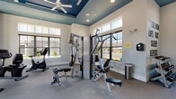 Located at 562 Washington Ave in Dumont, NJ, this fitness center is a thoughtfully designed, single-floor facility built to support a full range of workout routines. The space features a striking tray ceiling finished in a deep teal-blue, accented by recessed lighting and ceiling fans that keep the environment comfortable during intense sessions. Large, black-framed windows line multiple walls, flooding the interior with natural light and offering views of the surrounding neighborhood.

The cardio zone is well-stocked with multiple True-brand treadmills, elliptical machines, upright and recumbent stationary bikes, allowing users to choose from a variety of low- and high-impact options. A pair of wall-mounted flat-screen televisions provides entertainment throughout workouts. The strength training area includes a Hoist multi-station cable machine, a dedicated free weights rack with dumbbells, and a flat workout bench for versatile resistance training. Stability balls and additional accessories round out the functional fitness offerings.

Full-length mirrors along one wall expand the sense of space visually while allowing users to monitor their form. Motivational wall lettering — "Wake. Run. Lift. Eat. Sleep. Repeat." — adds an energetic touch to the overall ambiance. The flooring throughout is durable carpet suited for fitness use, and the neutral gray-and-white walls keep the space feeling clean, open, and professional.