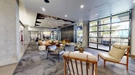 Located at 350 E Congress Street in downtown Tucson, Arizona, this contemporary communal lounge offers a generously scaled, single-floor open-plan layout. The space is defined by a striking mix of modern and industrial design elements that come together cohesively throughout.

At the heart of the lounge, a sleek built-in fireplace anchors a sophisticated seating area furnished with an eclectic mix of lounge chairs, sofas, and accent tables arranged atop patterned carpet. Overhead, a distinctive honeycomb-shaped pendant light installation adds a bold architectural focal point to the ceiling, complemented by recessed lighting throughout.

Floor-to-ceiling industrial-style black metal and glass garage doors line one side of the space, flooding the interior with natural light and offering direct visual and physical connection to an outdoor patio area with views of mature trees. This seamless indoor-outdoor relationship is a defining feature of the lounge.

Polished concrete flooring frames the perimeter and transitions into carpeted zones, while exposed concrete columns punctuate the space. Large-panel sliding barn doors with a textured stone finish provide flexible room division, leading to additional areas including a glass-enclosed conference room. Decorative wall art, including a historical photographic mural, adds character and a nod to Tucson's urban heritage. A game area with a foosball table is also visible, enhancing the space's versatility as a social hub.