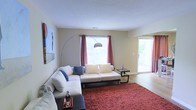 This single-story space centers around a well-appointed living room that is both functional and inviting. The room is bathed in natural light streaming through a large window dressed with rich red curtains, complemented by a sliding glass door that opens to an outdoor area, offering views of surrounding greenery and enhancing the sense of openness. The textured ceiling adds subtle architectural character overhead.

The living area is furnished with a generous white sectional sofa accented by blue throw pillows, anchored by a deep red shag area rug that introduces warmth and color contrast against the light hardwood-style flooring. A sleek arc floor lamp provides additional ambient lighting, while a dark-framed side table adds a modern touch. Wall art on the left side of the room contributes a creative focal point.

The neutral, beige-toned walls throughout create a versatile backdrop, allowing the furnishings and decor to stand out. The sliding door connects the interior to what appears to be a balcony or patio, expanding the functional living space outdoors. Overall, the space presents a clean, contemporary aesthetic with a comfortable and airy atmosphere.