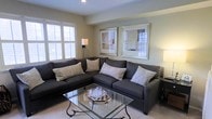 Located at 4604 Cascade Mills B, this single-floor unit offers a comfortable and stylishly designed living space. The living area serves as the focal point of the unit, showcasing a neutral color palette of soft beiges and warm grays that creates a calm, inviting atmosphere. A large, dark charcoal sectional sofa anchors the room, complemented by patterned accent pillows that add visual texture. A glass-top coffee table with an ornate metal base provides a sleek, contemporary touch while keeping the space feeling open and airy. Natural light filters in through windows fitted with white plantation shutters, offering both brightness and privacy control. The walls are adorned with framed artwork that reinforces the room's understated, sophisticated aesthetic. Warm accent lighting from floor and table lamps adds depth to the ambiance during evening hours. The light-toned flooring enhances the sense of openness throughout the space. Overall, the unit presents a well-balanced blend of comfort and refined design, making it a functional and visually appealing living environment.