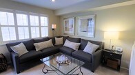 Located at 4604 Cascade Mills B, this single-floor unit offers a comfortable and stylishly designed living space. The living area serves as the focal point of the unit, showcasing a neutral color palette of soft beiges and warm grays that creates a calm, inviting atmosphere. A large, dark charcoal sectional sofa anchors the room, complemented by patterned accent pillows that add visual texture. A glass-top coffee table with an ornate metal base provides a sleek, contemporary touch while keeping the space feeling open and airy. Natural light filters in through windows fitted with white plantation shutters, offering both brightness and privacy control. The walls are adorned with framed artwork that reinforces the room's understated, sophisticated aesthetic. Warm accent lighting from floor and table lamps adds depth to the ambiance during evening hours. The light-toned flooring enhances the sense of openness throughout the space. Overall, the unit presents a well-balanced blend of comfort and refined design, making it a functional and visually appealing living environment.