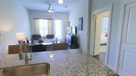 This single-floor residence is thoughtfully designed with an open-concept layout that seamlessly connects the kitchen and living areas. The kitchen features a generous granite countertop with subtle speckled patterning, offering ample prep and serving space, complemented by a stainless steel sink. The surface flows visually into the living room, creating a welcoming and connected atmosphere throughout.

The living area is well-proportioned, centered around a ceiling fan with integrated lighting that keeps the space comfortable and bright. Two windows dressed with light curtains framed by red accent valances allow natural light to fill the room. Seating is arranged around a coffee table, creating a relaxed and functional gathering space. A dark wood media console anchors the entertainment area, topped with a flat-screen television and decorative accents, including a tall ornamental grass plant in a woven basket.

An open doorway off the living space leads into a bedroom, which features a neatly made bed with colorful accent pillows, suggesting a calm and restful retreat. The overall palette of neutral grays and whites throughout gives the space a clean, cohesive, and modern feel.