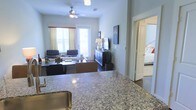 This single-floor residence is thoughtfully designed with an open-concept layout that seamlessly connects the kitchen and living areas. The kitchen features a generous granite countertop with subtle speckled patterning, offering ample prep and serving space, complemented by a stainless steel sink. The surface flows visually into the living room, creating a welcoming and connected atmosphere throughout.

The living area is well-proportioned, centered around a ceiling fan with integrated lighting that keeps the space comfortable and bright. Two windows dressed with light curtains framed by red accent valances allow natural light to fill the room. Seating is arranged around a coffee table, creating a relaxed and functional gathering space. A dark wood media console anchors the entertainment area, topped with a flat-screen television and decorative accents, including a tall ornamental grass plant in a woven basket.

An open doorway off the living space leads into a bedroom, which features a neatly made bed with colorful accent pillows, suggesting a calm and restful retreat. The overall palette of neutral grays and whites throughout gives the space a clean, cohesive, and modern feel.