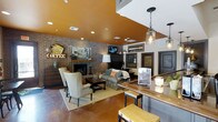 This single-floor space exudes a warm, inviting atmosphere through a thoughtfully curated blend of industrial and rustic design elements. The open-concept layout seamlessly connects a cozy lounge area with a bar or counter seating section, creating a versatile environment ideal for socializing and relaxing.

The lounge area is anchored by a brick fireplace, which serves as a striking focal point against the exposed brick accent wall. Comfortable seating, including classic wingback chairs and an upholstered sofa, is arranged around a wooden coffee table atop a neutral area rug, fostering an intimate gathering space. Decorative touches such as vintage-style marquee signage and eclectic wall art add personality and character throughout.

Overhead, a bold warm amber-painted ceiling draws the eye upward and adds a vibrant, energetic contrast to the earthy tones of the brick and polished concrete flooring below. Pendant lighting with mason jar-style fixtures illuminates the bar counter area, which features a rich wood-top surface and industrial-style metal stools.

The polished concrete floors tie the entire space together, reinforcing the industrial aesthetic while remaining practical and durable. Overall, this space strikes a balance between rustic charm and modern edge, making it a distinctive and memorable environment.