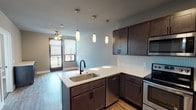 This single-floor unit offers a thoughtfully designed open-concept layout that seamlessly connects the kitchen and living area. The kitchen is a standout feature, showcasing rich dark-brown cabinetry, crisp white countertops, and a classic white subway tile backsplash. A peninsula with an undermount stainless steel sink and matte black faucet serves as both a functional workspace and a casual gathering point, illuminated by a trio of elegant pendant lights overhead. The kitchen is fully equipped with stainless steel appliances, including a range, microwave, dishwasher, and refrigerator, all set atop warm-toned hardwood-style flooring. The adjacent living area benefits from natural light streaming through large windows dressed with blinds, and is finished with a ceiling fan. The bathroom continues the refined aesthetic with dark cabinetry, white countertops, matte black fixtures, and a large mirror with overhead lighting. A convenient in-unit washer and dryer is accessible directly from the bathroom. The bedroom offers plush carpeting, a ceiling fan, a sliding-door closet with built-in shelving, and a window that brings in natural light, creating a comfortable and restful retreat.