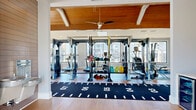 Located at Fountain Grove Circle in Fayetteville, NC, this fitness center offers a comprehensive workout environment across a single, generously sized floor. The main workout area is an open, light-filled space anchored by a striking vaulted wood-plank ceiling with exposed white beams and ceiling fans, lending a warm and upscale aesthetic. Large windows and a full-panel glass garage door flood the floor with natural light and provide views of the surrounding community. Light hardwood-style flooring runs throughout, complementing the bright, neutral walls. The main floor houses an extensive array of commercial-grade equipment, including multi-functional cable machines, power racks, treadmills, ellipticals, rowing machines, benches, and free weights. A long turf sprint track runner with yardage markings anchors the center of the floor, ideal for functional training and speed work. A glass-enclosed studio space offers stationary bikes and additional strength equipment alongside a wall-mounted TV, providing a focused environment for group or individual training. The facility also includes a utility room and a dedicated bathroom for added convenience. With its architectural detail, abundant natural light, and comprehensive equipment selection, this fitness center delivers both a functional and inspiring training experience.