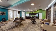 Located at 7107 Brandy Run Dr in Mechanicsville, Virginia, this single-story community amenity center is designed to serve as a dynamic hub for recreation, wellness, and productivity. The space is immediately distinguished by its bold, energetic color palette—teal, lime green, and yellow accents appear throughout, creating a lively and modern atmosphere.

The entry area features a reception desk framed by a wood-paneled tray ceiling with teal accents and curated artwork, setting a welcoming tone. Adjacent to it, a spacious game lounge hosts a pool table, foosball table, and comfortable seating, all beneath sculptural ceiling details and recessed lighting.

The full kitchen is a standout feature, boasting dark espresso cabinetry, a long quartz-topped island with bar seating, stainless steel appliances, and a striking green accent wall beneath an ornate circular ceiling installation.

The fitness center is well-equipped with cardio machines including ellipticals and treadmills, a multi-use cable machine, free weights, and floor-to-ceiling mirrors, all on durable rubber flooring with bold yellow and teal painted walls adding visual energy.

Two conference rooms offer professional settings with long tables, ergonomic seating, wall-mounted displays, and linear pendant lighting. A separate lounge area with vibrant upholstered seating and patterned carpeting provides an informal gathering space throughout.