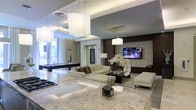 This single-floor space showcases a refined, contemporary design defined by an open-plan layout that connects multiple functional zones in one expansive area. The flooring is clad in polished marble, lending a bright, high-end aesthetic that extends throughout the space. Overhead, a layered ceiling with recessed lighting and cove detailing adds architectural depth, while oversized pendant light fixtures — featuring soft, diffused shades — serve as striking focal points that anchor each zone.

The living area is furnished with a plush, cream sectional sofa accented with textured throw pillows, positioned around a dark-framed coffee table and oriented toward a wall-mounted flat-screen television. The media wall features rich dark wood paneling flanked by a textured stone or mosaic tile accent, adding visual contrast to the otherwise light-toned palette. A white upholstered ottoman completes the seating arrangement, reinforcing the cohesive neutral color scheme.

Natural light enters generously from floor-to-ceiling windows visible toward the rear of the space. A billiards table is positioned in the background, indicating a dedicated recreation zone. A white orchid centerpiece on the marble surface adds an elegant, organic touch to the overall ambiance.