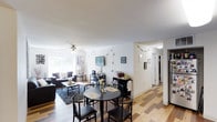 This single-floor space offers a welcoming and functional open-concept layout that seamlessly connects the dining and living areas. The flooring throughout is a warm hardwood-style material that adds a sense of continuity and warmth to the space. A round black dining table with matching chairs anchors the dining area, positioned conveniently near the kitchen access, making it ideal for everyday meals and casual gatherings. A textured dark area rug defines the living room zone, where comfortable seating is arranged around a central coffee table. A wall-mounted flat-screen television serves as a focal point, flanked by decorative accents that add personality to the space. A ceiling fan with a light fixture overhead provides both ventilation and ambient lighting in the main living area. Large windows along one wall invite an abundance of natural light, enhancing the airy and open feel of the room. A compact storage and utility area along one side offers additional organization options. The entryway corridor, visible from the main living space, leads to additional rooms, providing a clear and efficient flow throughout the home. The overall ambiance is casual, bright, and livable.
