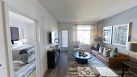 This single-floor residence offers a thoughtfully designed living space that balances comfort and modern style. The main living area is bathed in natural light, courtesy of large windows dressed with sheer curtains that allow sunlight to filter through while maintaining a sense of openness. A glazed door leads directly to an outdoor balcony, seamlessly blending indoor and outdoor living.

The living room is anchored by rich hardwood-style flooring and features a tasteful arrangement of furnishings, including a leather sofa, an accent armchair, and a round coffee table set atop a layered area rug. A wall-mounted flat-screen television sits above a dark storage credenza, adding both function and visual interest to the space. Warm, neutral wall tones are complemented by subtle gray accents, creating a calm and inviting atmosphere throughout.

An adjacent bedroom is partially visible, hinting at well-organized closet storage and coordinated bedding. The overall layout feels efficient yet spacious, with clean lines and contemporary décor choices that lend the home a polished, move-in-ready appeal. Natural light, modern finishes, and a cohesive design palette make this a welcoming and practical living environment.