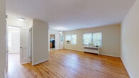 This single-floor residence is thoughtfully laid out across a well-proportioned floor plan, as revealed by the 3D scan. Warm-toned hardwood flooring runs throughout the main living areas and bedrooms, contributing a sense of continuity and comfort to the space.

The living area is generously sized, featuring multiple windows that frame views of surrounding greenery and allow abundant natural light to fill the room. A pendant light fixture overhead adds a classic touch, while a radiator and window-mounted air conditioning unit provide year-round climate control. A wall-mounted mini-split unit is also present, offering supplemental temperature management.

The kitchen is equipped with warm wood cabinetry, granite-style countertops, a black gas range, a full-size refrigerator, and a sink. Tile flooring and a window with outdoor views complete the practical and functional kitchen space. A closet is conveniently located adjacent to the kitchen area.

The bedroom is a comfortable, well-proportioned room with the same warm hardwood flooring, a window with natural light, and a radiator beneath it for efficient heating.

The bathroom features classic white tile wainscoting, tile flooring, a full bathtub with a shower and curtain, and a toilet, presenting a clean and straightforward layout.