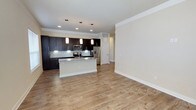 This single-floor space showcases a contemporary open-concept layout that seamlessly connects the kitchen and living areas. The kitchen is a standout feature, equipped with dark espresso cabinetry that contrasts elegantly against a classic white subway tile backsplash. A central island with a granite countertop serves as both a functional workspace and a natural gathering point, illuminated by a trio of modern pendant lights overhead. Stainless steel appliances, including a French door refrigerator and a built-in microwave, add a polished, professional finish to the culinary space.

Recessed lighting complements the pendant fixtures, ensuring the kitchen is well-lit throughout. Light-toned hardwood flooring extends across the entire open area, lending warmth and a sense of continuity to the space. The expansive living area benefits from neutral wall tones and crisp white crown molding and baseboards, creating a clean, refined aesthetic. A hallway with a white door leads to additional areas of the unit. The overall ambiance is modern, bright, and welcoming, with a cohesive design that balances style and functionality.