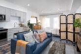This thoughtfully designed single-floor studio offers an open-concept layout that maximizes functionality and flow. Light-toned hardwood-style flooring runs throughout, tying together each area of the space cohesively.

The living area serves as the heart of the studio, featuring recessed lighting and a ceiling fan overhead. A bold abstract artwork anchors one wall above a richly grained credenza, while a navy leather sofa, woven ottoman, and layered area rugs lend warmth and character. A rattan room divider provides a natural visual separation between the living and sleeping zones.

The sleeping area is bathed in natural light from large windows dressed with patterned curtains. A mid-century modern dresser, vibrant wall art, and lush potted plants complete the serene and well-curated aesthetic.

The open kitchen is fitted with full-height grey shaker cabinetry, stainless steel appliances including a French door refrigerator, electric range with over-the-range microwave, and dishwasher. A subway tile backsplash and clean white countertops add a crisp, modern finish.

A walk-in closet off the main living area offers ample storage, while the bathroom features a white undermount vanity, grey cabinetry with open shelving, and a large mirror, creating a bright and functional space.