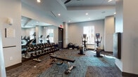 This fitness center in Jacksonville, Florida, offers a comprehensive workout environment designed to accommodate a wide range of exercise routines. The space is thoughtfully laid out across a single floor, with distinct zones dedicated to cardio training, strength machines, and free weights. The interior features a modern aesthetic, with neutral gray walls, recessed lighting, and patterned carpet tiles in a mix of gray and teal tones that add visual interest underfoot. Ceiling fans provide additional air circulation throughout the space.

The cardio zone is well-appointed with multiple treadmills, elliptical trainers, and stair climbers, all positioned near windows that allow natural light to filter in and offer views of the surrounding area. A large floor-to-ceiling mirror along one wall visually expands the space and is especially useful for the free weight and functional training area, which includes a full rack of dumbbells, a workout bench, and stability balls. 

The strength training section houses a variety of selectorized machines, including leg press, leg curl, and cable systems, with wall-mounted flat-screen televisions positioned nearby. A rowing machine rounds out the available equipment. The overall ambiance is clean, functional, and modern.