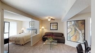 This single-floor unit offers an open, efficiently designed layout that seamlessly blends the living and sleeping areas into one cohesive space. A distinctive white-painted partial divider with vertical slat detailing provides a soft visual separation between the sleeping area and the living room, maintaining an airy, open feel while offering a subtle sense of privacy. The sleeping area is furnished with a full-sized bed and complemented by a bedside lamp, while framed artwork adorns the walls throughout, adding a personal and artistic touch to the space. The living area features a plush sofa, a sleek glass-top coffee table, and a ceiling fan with a light fixture overhead, ensuring both comfort and functionality. Textured popcorn ceilings and warm beige carpeting run throughout, creating a consistent and inviting ambiance. The neutral wall tones are accented by carefully placed floor lamps that cast a warm, welcoming glow. Decorative elements, including a vase of fresh flowers and multiple framed art pieces, contribute to the lived-in and tastefully curated character of the space. Overall, the unit offers a practical and comfortable environment with a warm, residential atmosphere.