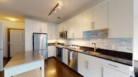 This well-appointed single-floor residence offers a thoughtfully designed layout that balances modern aesthetics with practical functionality. The kitchen is a standout feature, showcasing crisp white cabinetry extending to the ceiling, dark granite countertops, and a textured stone tile backsplash that adds visual depth. A full suite of stainless steel appliances — including a refrigerator, range, over-the-range microwave, and dishwasher — is complemented by track lighting overhead and under-cabinet illumination. Rich hardwood flooring runs throughout the kitchen and connects seamlessly into the open living area, which is bright and airy thanks to multiple windows with blinds allowing natural light to fill the space. A glass-panel door in the living area provides additional light and potential access to the outdoors. The bedroom features soft carpeting and a window with a view of the surrounding greenery, offering a quiet, comfortable retreat. The bathroom is finished with neutral tile flooring, a white soaking tub and shower combo, a clean white countertop vanity, and dark espresso cabinetry for contrast. An in-unit laundry area adds further convenience to this move-in-ready space.