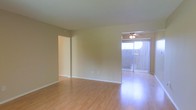This single-floor space features an open living area finished with smooth hardwood-style flooring that reflects natural light throughout the room. The warm, neutral-toned walls create a clean and inviting atmosphere, complementing the bright, airy feel of the space. A ceiling fan with an integrated light fixture is positioned near the sliding glass door, providing both ventilation and additional illumination. The sliding glass door is dressed with vertical blinds and opens to a private outdoor balcony or patio area, allowing natural light to fill the interior while offering views of the exterior surroundings. A doorway on the adjacent wall leads to a separate room or hallway, suggesting a functional layout with distinct living zones. The white baseboards throughout provide a crisp contrast to the flooring and walls, adding a polished finish to the overall aesthetic. The open layout and abundant natural light make this a practical and comfortable living space.