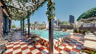 Located at 808 Constance St in the heart of New Orleans, this stunning rooftop pool deck offers a sophisticated outdoor retreat with striking visual character. The centerpiece is a generously sized rectangular lap pool with crystal-clear turquoise water, framed by a bold red-and-white checkered tile surround that adds a playful yet refined aesthetic. Exposed brick facades line the perimeter of the rooftop, adorned with climbing vines and lush greenery that cascade from pergola structures overhead, creating a seamless blend of natural and architectural elements. Shade is provided by a row of distinctive green-and-white tropical print umbrellas, complemented by cushioned lounge chairs and daybeds positioned for optimal sun and skyline exposure. Café-style bistro tables and chairs are tucked beneath the vine-draped pergola, offering a more intimate setting for dining or relaxing. Large industrial-style windows and glass doors along the brick building facade allow for an indoor-outdoor connection, enhancing the open-air ambiance. The rooftop commands panoramic views of the New Orleans downtown skyline, with clear sightlines to surrounding city landmarks. The overall atmosphere is a dynamic fusion of Southern charm, resort-style luxury, and urban energy, making it a distinctive destination in the city.