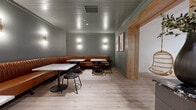 Located at 7510 N Oak Trafficway in Gladstone, Missouri, this intimate dining space offers a polished and cohesive interior design that balances modern sophistication with warm, inviting elements. The room is defined by a continuous channel-tufted cognac leather banquette that lines the walls, providing generous seating capacity while serving as a striking visual anchor throughout the space.

Each seating station is outfitted with marble-top tables featuring distinctive grey and white veining, paired with sleek black metal chairs that complement the room's refined aesthetic. Warm-toned gold wall sconces are evenly distributed along the sage green walls, casting a soft, ambient glow that enhances the intimate atmosphere of the dining environment.

The ceiling features a painted tongue-and-groove paneled treatment, adding textural depth overhead, while light-toned hardwood flooring runs throughout, contributing to the airy and welcoming feel underfoot. A textured tile accent wall lines one side of the space near a dark cabinetry credenza with a granite countertop, providing both functional storage and a sophisticated design contrast.

A natural wood-framed doorway connects this dining area to an adjacent space, and framed artwork is thoughtfully placed to add character. The overall ambiance is one of curated elegance, well-suited for a boutique dining or hospitality experience.