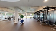 Located at 1641 Davie St in Vancouver's West End, this two-floor fitness facility — branded "HOUSE" — offers a well-appointed and generously sized exercise environment spread across two distinct levels. The larger main floor features an open, light-filled layout with floor-to-ceiling windows that frame views of a manicured outdoor green space, flooding the room with natural light. Structural white columns punctuate the floor plan, adding an architectural rhythm to the space. This level is equipped with a comprehensive free weights area, including barbells, plate-loaded racks, dumbbells, kettlebells, and medicine balls organized on sleek multi-tiered storage units. Full-length mirrors line the walls, enhancing the sense of openness. A curved staircase with a sculptural design connects the two floors, serving as both a functional and aesthetic centerpiece. The upper floor continues the bright, airy aesthetic with large windows offering urban and treetop views. This level houses an array of cardio equipment, including treadmills, ellipticals, and stationary bikes, all positioned to take advantage of the natural light and outdoor sightlines. Throughout both floors, durable rubber flooring, linear LED lighting, and exposed ceiling elements contribute to a clean, modern, and professional atmosphere.