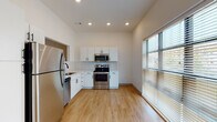 This single-floor unit presents a clean, contemporary design highlighted by a cohesive neutral palette and quality finishes throughout. The kitchen serves as a central feature, outfitted with stainless steel appliances including a refrigerator, electric range, built-in microwave, and dishwasher. White shaker-style cabinetry with matte black hardware lines the walls, complemented by a white subway tile backsplash and light-toned countertops. A stainless steel sink with a matte black faucet anchors the counter space, and a pass-through window connects the kitchen to an adjoining room, enhancing openness. Light wood-look flooring extends through the kitchen and main living areas, adding warmth to the modern aesthetic. Recessed lighting provides consistent illumination throughout, while large windows fitted with horizontal blinds allow natural light to fill the space. The bedroom features neutral carpet and a window, creating a quiet, comfortable retreat. The bathroom is finished with a single-sink vanity topped with a white countertop, a large mirror, brushed nickel fixtures, a tiled walk-in shower with glass door, and gold-toned accent hardware. Overall, the unit reflects a well-considered, move-in-ready design with modern functionality.