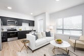 This single-floor residence offers a well-designed open-concept layout that seamlessly integrates the living, dining, and kitchen areas. The living room is bathed in natural light from multiple large windows with horizontal blinds, offering views of surrounding greenery. A standout textured concrete-style accent wall anchors the space and provides a striking backdrop for the entertainment area, complementing the warm light wood flooring found throughout.

The kitchen is outfitted with dark matte cabinetry contrasted by crisp white countertops and a classic subway tile backsplash. Stainless steel appliances — including a refrigerator, electric range, built-in microwave, and dishwasher — provide a full suite of modern conveniences, while a deep stainless steel sink completes the functional workspace.

The bedroom is a calm, neutral retreat featuring warm-toned walls and ample room for furnishings, with a closet for storage. The bathroom is clean and bright, finished with light wood flooring and a shower-tub combination. A stacked in-unit washer and dryer is conveniently integrated into the space, adding practicality without sacrificing footprint. The overall ambiance is modern, light-filled, and thoughtfully designed.