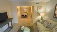 This single-floor residence offers a thoughtfully designed open-concept layout that seamlessly connects the living and dining areas. The living room provides a welcoming atmosphere, highlighted by neutral beige carpeting, soft ambient lighting from a stylish table lamp, and decorative wall art featuring floral motifs that add a touch of warmth and color. A plush sofa and a dark-toned area rug anchor the space, while a round accent table completes the seating arrangement.

A wide archway leads directly into the dining room, which is distinguished by a rich olive-green accent wall that creates a striking contrast against the otherwise neutral palette. The dining area features a wooden table surrounded by matching chairs, centered beneath a contemporary pendant chandelier that provides focused, elegant lighting. A framed piece of artwork adorns the accent wall, enhancing the room's refined character.

Throughout both spaces, the consistent carpeting and neutral wall tones create a cohesive, harmonious flow. The ceiling-mounted HVAC vent indicates a central air system, contributing to everyday comfort. Overall, the space exudes a calm, inviting ambiance well-suited for both relaxed living and formal dining.
