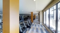 This fitness center offers a spacious, well-appointed workout environment designed to accommodate a range of exercise needs. The room is anchored by a row of treadmills and cardio machines aligned along one side, providing ample equipment for cardiovascular training. Full-length mirrored walls extend the visual depth of the space, creating an open and expansive feel while allowing users to monitor their form during workouts. Structural columns finished in a warm tan tone punctuate the room at regular intervals, contributing to the overall architectural character. The flooring features a distinctive patterned carpet tile in a combination of gray, blue, and tan tones, adding a modern, dynamic quality underfoot. Recessed lighting is evenly distributed across the ceiling, ensuring consistent illumination throughout the space. Large windows along one wall invite natural light and offer views to the exterior, enhancing the overall ambiance. Toward the rear of the room, additional equipment is visible, suggesting a comprehensive selection of fitness options. The corridor visible beyond hints at further amenities nearby, making this fitness center part of a larger, well-considered facility.