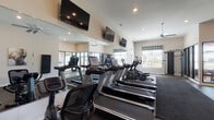 Located at 950 Brushfield Dr in Galloway, Ohio, this fitness center is a thoughtfully designed, single-floor facility built to support a comprehensive workout experience. The space features durable rubberized flooring throughout, providing both safety and comfort underfoot, with area rugs designating specific workout zones. Elevated ceilings contribute to an open, airy atmosphere, while ceiling fans and recessed lighting ensure the environment remains comfortable and well-lit. Multiple flat-screen televisions are mounted along the walls, offering entertainment during cardio sessions. A full wall of mirrors amplifies the sense of space and allows users to monitor their form. The cardio zone is equipped with a row of treadmills and elliptical machines, complemented by a stationary bike. A dedicated strength training area includes a multi-function cable machine, an adjustable weight bench, and a full rack of dumbbells ranging in various sizes. Motivational fitness artwork is displayed along the walls, adding an energizing aesthetic. Large windows allow natural light to flow in, offering views of the surrounding outdoor area. Glass partition walls connect the fitness center to an adjacent interior space, providing visual openness. An exterior door offers convenient direct access from outside, rounding out this functional and well-appointed facility.