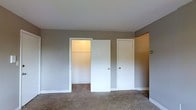 This single-floor space includes a room finished in neutral taupe-toned walls and white trim, creating a clean and understated aesthetic. The flooring is wall-to-wall carpet in a warm beige tone, contributing to a comfortable and quiet atmosphere. Two white-painted doors are positioned along one wall — one leads to a built-in closet, which is open and reveals interior shelving illuminated by a soft warm light, offering organized storage potential. The second door, fitted with a standard knob, likely leads to an adjoining room or hallway. The white baseboards provide a crisp contrast against the neutral walls, while the smooth, white ceiling completes the simple, functional design. The overall ambiance is calm and neutral, well-suited for use as a bedroom or private living space. The layout appears straightforward with no decorative embellishments, emphasizing practicality. The closet's built-in shelving adds a notable storage feature, making this room functional for everyday living.