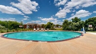 Located at 1828 Sandy Point Rd in Bryan, Texas, this generously sized outdoor amenity area offers a well-appointed recreational experience across a single-level layout. The centerpiece of the space is a large, kidney-shaped swimming pool with vivid turquoise water, framed by a wide concrete pool deck finished with brick-tiled edging. Multiple modern lounge chairs are positioned along the deck, providing ample space for sunbathing and relaxation under the open Texas sky.

Adjacent to the pool, a substantial white pergola structure offers shaded outdoor seating with tables and chairs, making it an ideal gathering spot for residents seeking relief from the sun. A cornhole board and additional lounge seating are also present, enhancing the recreational appeal of the area. The surrounding landscape features mature trees and lush greenery that provide a natural, park-like backdrop, while a black wrought-iron perimeter fence encloses the entire area for safety and security.

The brick-accented building visible in the background reflects a traditional architectural style consistent with the broader residential community. With open views of the sky and surrounding landscape, the space maintains a bright, airy ambiance throughout, making it a versatile and inviting outdoor destination well-suited for both relaxation and social activity.