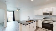 Located at 1200 E 15th St in Plano, TX, this one-bedroom, one-bathroom unit offers a thoughtfully designed layout with modern finishes throughout. Light wood-style flooring runs seamlessly across the entire space, creating a bright and cohesive aesthetic.

The kitchen is a standout feature, showcasing white shaker-style cabinetry, dark countertops, a light blue subway tile backsplash, and a full suite of stainless steel appliances including a French door refrigerator, range with microwave above, and dishwasher. A peninsula with an integrated sink provides additional prep space and a natural division between the kitchen and living area. Recessed lighting illuminates the space effectively.

The bedroom is a comfortable, light-filled room with a ceiling fan and a window offering views of surrounding greenery. A closet area within the bedroom conveniently houses a stacked washer and dryer unit, adding practical in-unit laundry functionality.

The bathroom is clean and well-appointed, featuring white subway tile surrounding a full soaking tub and shower combination, a white vanity with dark countertop, and the same light wood-style flooring carried throughout the unit. The overall design palette — whites, light wood tones, and dark countertop accents — gives the space a fresh, contemporary feel.