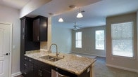 This single-floor space showcases an open-concept layout that connects the kitchen and living area in a cohesive, functional design. The kitchen is appointed with rich dark cabinetry that provides a striking contrast against the light granite countertops, which extend across a peninsula-style island complete with a double stainless steel sink and a sleek pull-down faucet. Pendant lighting hangs above the island, adding focused illumination and a decorative touch. A tile backsplash accents the cooking area, complementing the overall aesthetic. A dishwasher is seamlessly integrated beneath the counter. The kitchen transitions naturally into the adjoining living area, which benefits from two windows fitted with white horizontal blinds, allowing ample natural light to fill the space. Neutral-toned walls and light carpeting in the living area create a calm, welcoming ambiance. A ceiling fan is centrally positioned overhead, providing both air circulation and an additional light source. The open flow between the kitchen and living area makes this space well-suited for everyday living and casual entertaining.