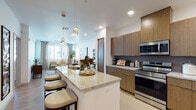 This well-appointed single-floor residence showcases a contemporary design with a cohesive blend of warm wood tones, granite surfaces, and light hardwood flooring throughout. The open-concept kitchen is a standout feature, equipped with full-height flat-front cabinetry in a rich wood finish, a textured tile backsplash, stainless steel appliances including a range, microwave, refrigerator, and dishwasher, and an extended granite-topped island with bar seating and pendant lighting overhead. The kitchen flows seamlessly into the dining and living areas, creating an expansive, connected layout ideal for entertaining. The living room is bright and welcoming, with multiple windows dressed in patterned drapes that invite natural light while offering privacy, complemented by a ceiling fan overhead. Two bedrooms provide comfortable retreats, each with plush carpeting, ceiling fans, and windows with window treatments. The primary bathroom features a lengthy dual-sink granite vanity with wood cabinetry, a large mirror with overhead lighting, and direct access to the primary bedroom. Clean white walls, recessed lighting, and consistent material choices throughout contribute to a polished, modern ambiance.